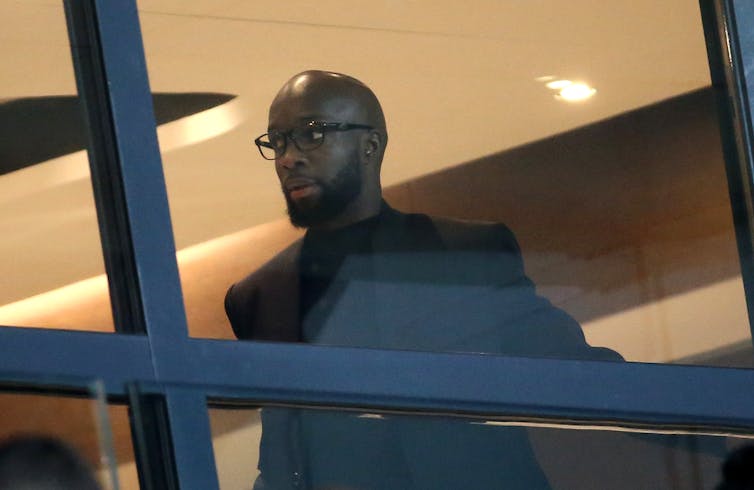 A man in a black suit and glasses looks out a window