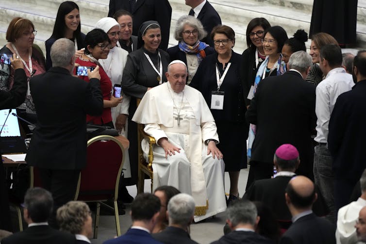 A group of women stands, many smiling, around a seated man in white robes with a cross around his neck.