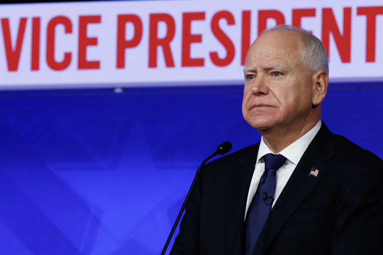 A balding man in a dark suit in front of a microphone looking intense.