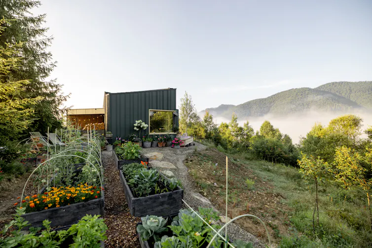 A vegetable garden sits outside a small, box-shaped home. In the distance rise a few hills that are shrouded in mist