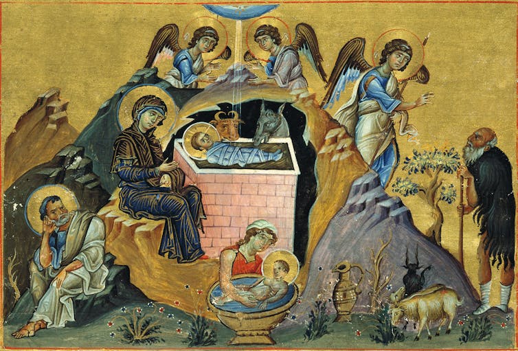 A woman sits with a swaddled baby in a manger as angels and animals look on.