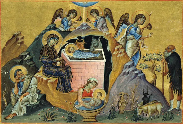 A woman sits with a swaddled baby in a manger as angels and animals look on.