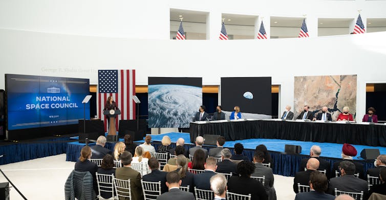 A group of people gathered around a large table, with Kamala Harris standing at a podium at the front next to a screen that says 'National Space Council.'