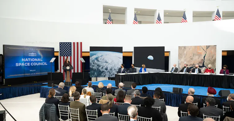 A group of people gathered around a large table, with Kamala Harris standing at a podium at the front next to a screen that says 'National Space Council.'