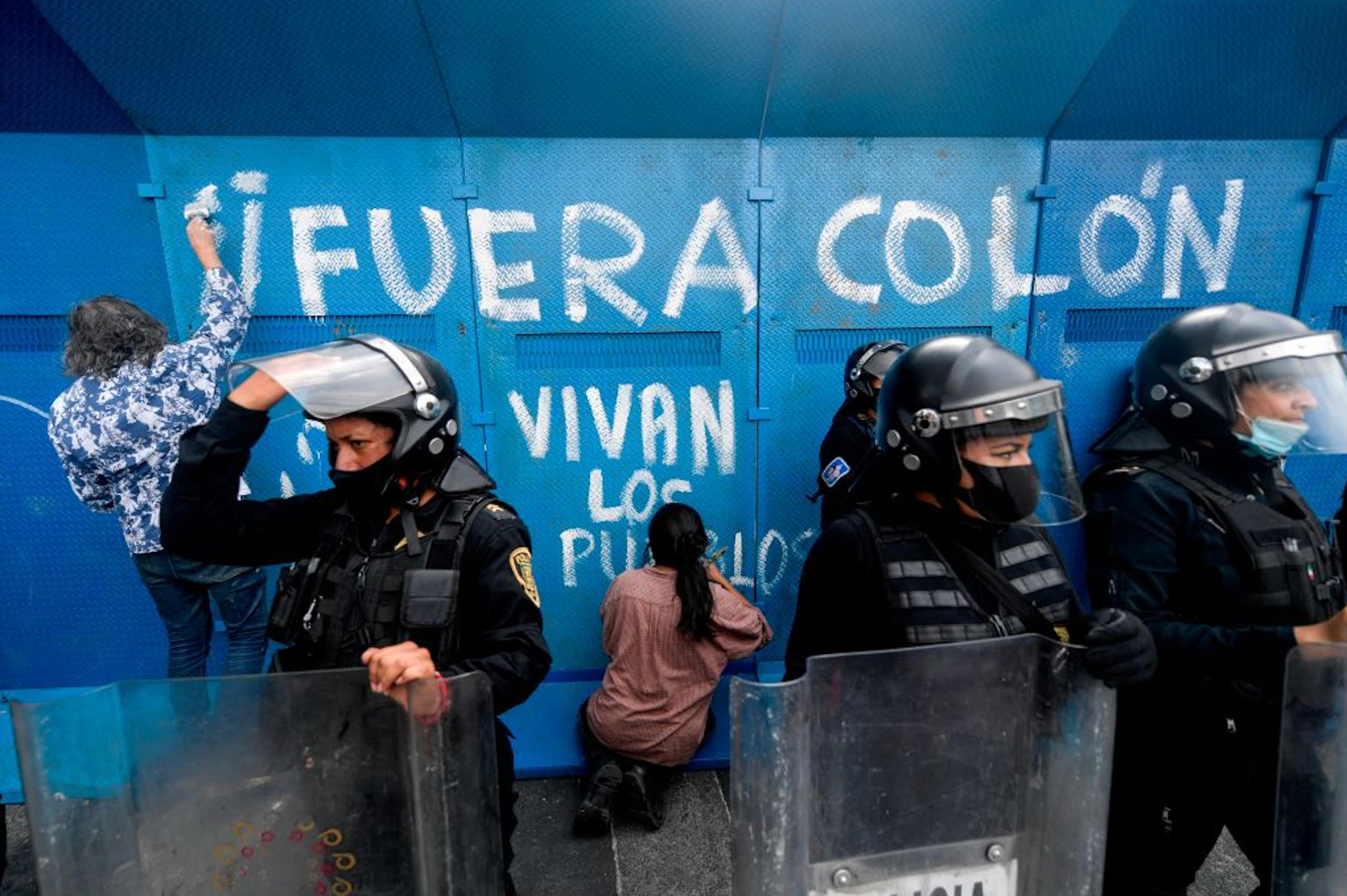 Two people with their backs to the camera write on a blue surface with white paint, as riot police stand nearby.