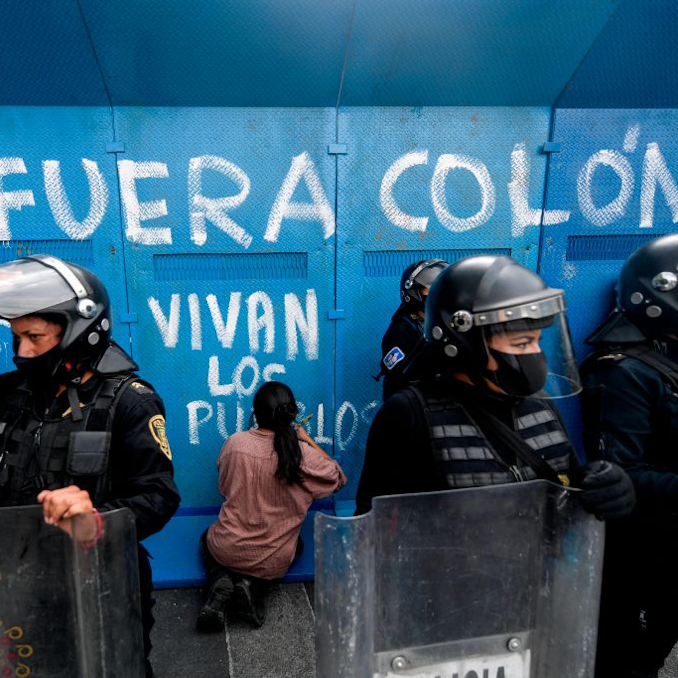 Two people with their backs to the camera write on a blue surface with white paint, as riot police stand nearby.