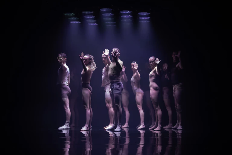 group of dancers stand under spotlight on stage, hands palm first to camera