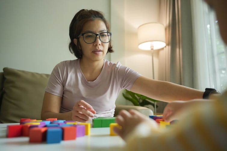 A woman reaches for a block on a table. A child is on the other side.