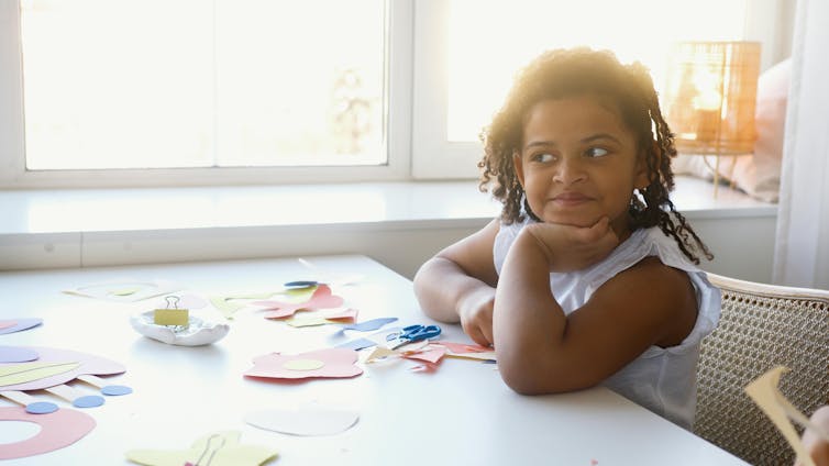 A young girl rests her chin in her hand. There are scissors and coloured papers on a table.