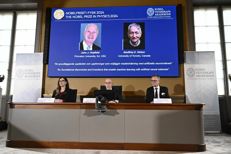 Photo showing three people giving a press conference at a desk. Behind them is a screen showing two men described as John Hopfield and Geoffrey Hinton