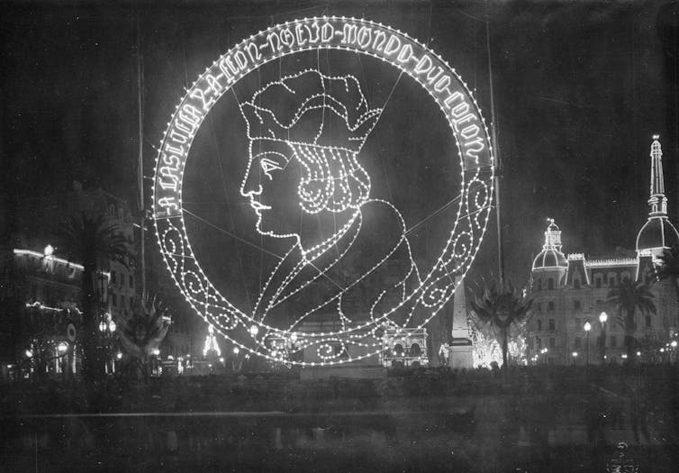 A huge image of Christopher Colombus, made up of strings of lights, towers over a plaza at night with buildings in the background.
