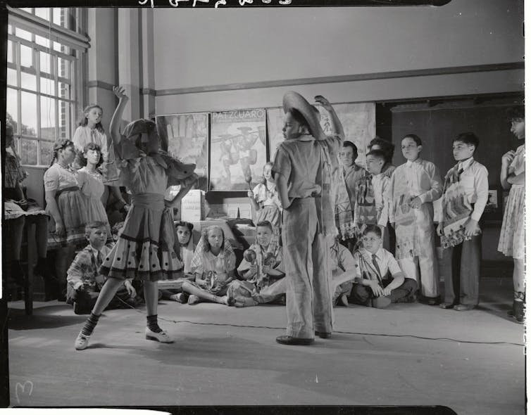 Children gather around a boy in a sombrero and a girl in a dress who are posing dramatically as they dance in a classroom.