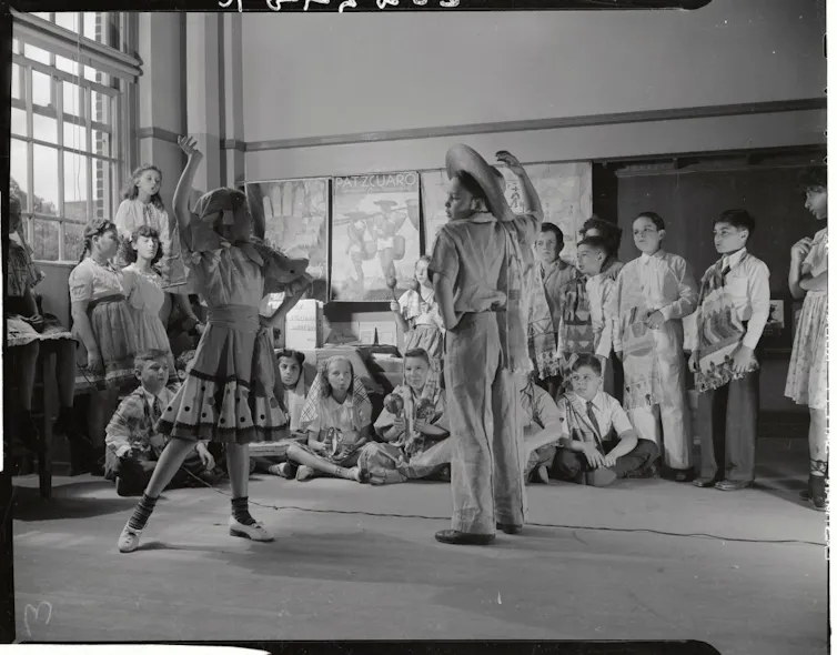 Children gather around a boy in a sombrero and a girl in a dress who are posing dramatically as they dance in a classroom.