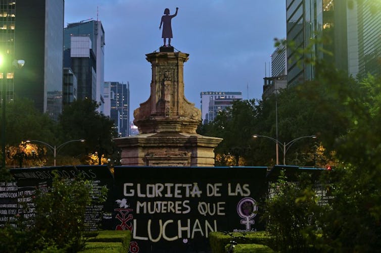 At dusk, a view of a statue of a woman holding up her arm, atop a high pedestal, with skyscrapers in the background.