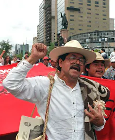 A man in a white shirt, with a mustache and broad-brimmed hat, raises his hand during a demonstration.