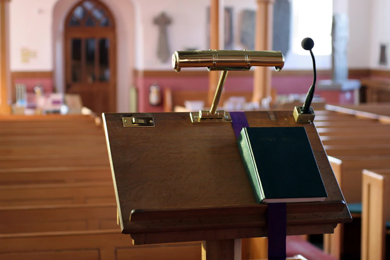 A Bible sits on a lectern with a small microphone and light on it, in front of rows of church pews.