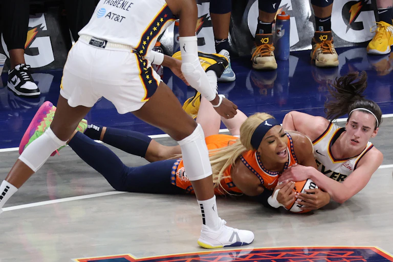 Two young women in basketball jerseys on their stomachs fighting for a basketball.