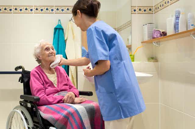 White-haired woman sitting in a wheelchair in a bathroom looking up at a dark-haired woman as she helps with face washing