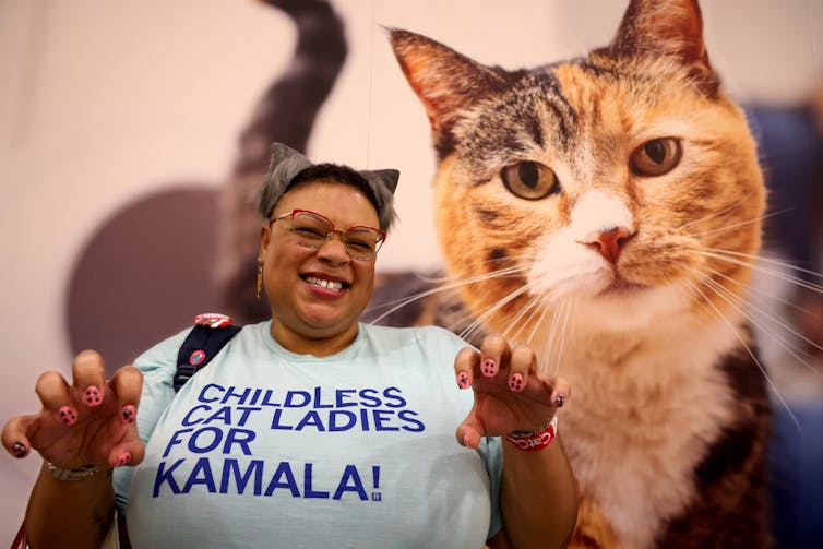 A woman wears a blue shirt that says 'Childless cat ladies for Kamala!' and stands in front of a large picture of a cat. She holds her hands out like claws.