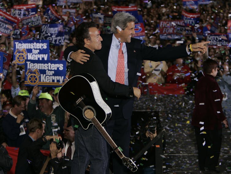 In front of a large crowd, two men -- one with a guitar -- stand close together, one with an arm around the other.
