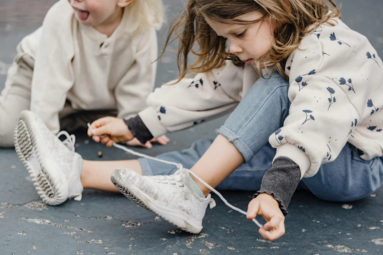 A young girl sits on the ground, trying to tie her shoelace.
