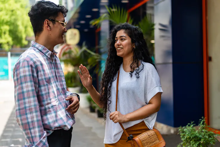A young couple chat to each other in the street. He is wearing a cotton long sleeve button up shirt, she is wearing a light coloured top.