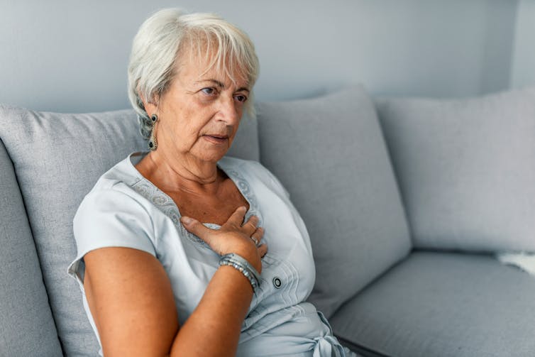 A woman sits on her couch feeling a bit breathless and clasping at her chest.