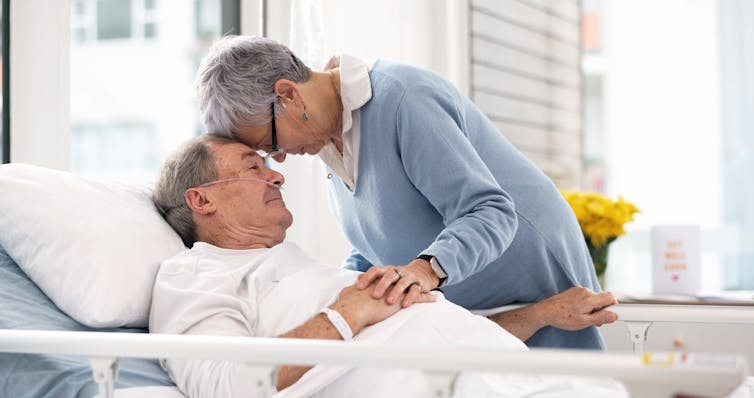A couple bend their heads together in hospital. The man is lying down, and he has a breathing support apparatus attached to his nose.