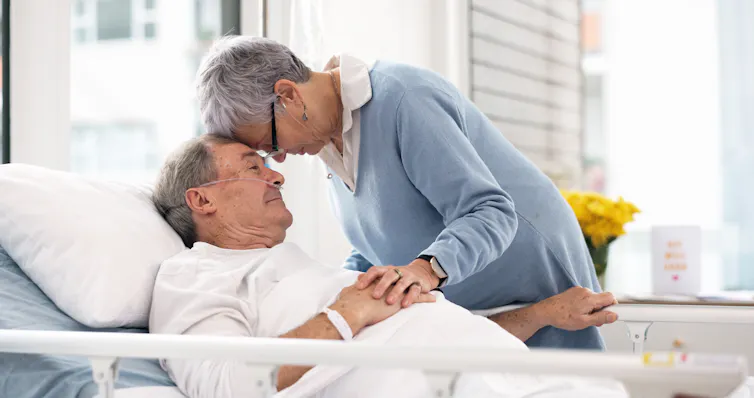 A couple bend their heads together in hospital. The man is lying down, and he has a breathing support apparatus attached to his nose.