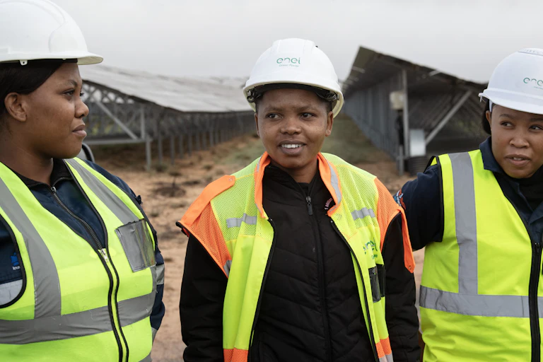 Three young women wearing yellow high visibility vests and white hard hats stand in front of rows of solar panels