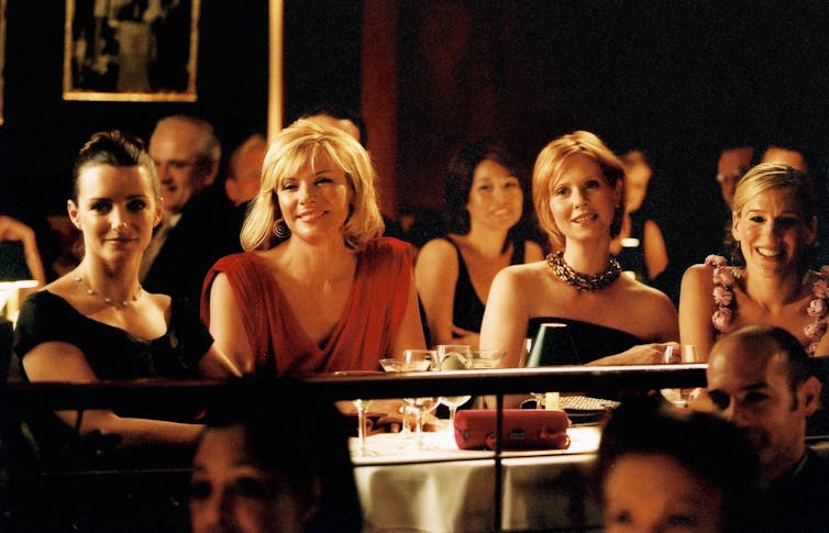 Still from Sex and the City, the four main characters sitting next to each other at a table in a restaurant and smiling