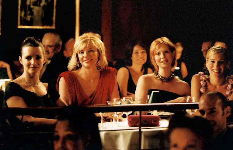 Still from Sex and the City, the four main characters sitting next to each other at a table in a restaurant and smiling