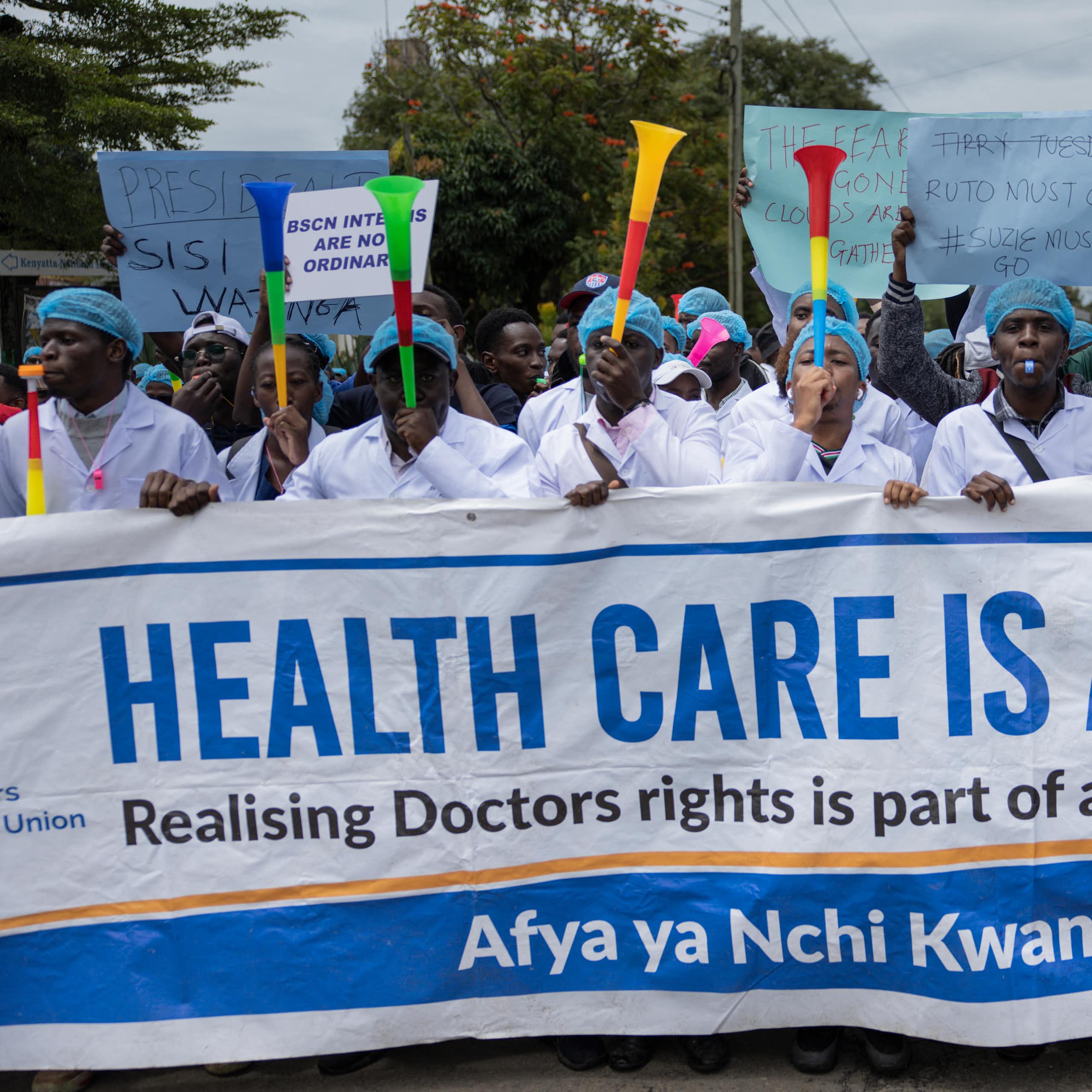 A crowd of people blowing vuvuzelas and holding up a placard written 'Healthcare is a right'