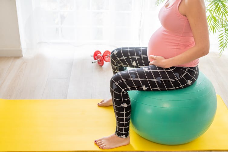 A pregnant woman sits on an exercise ball on a yoga mat.