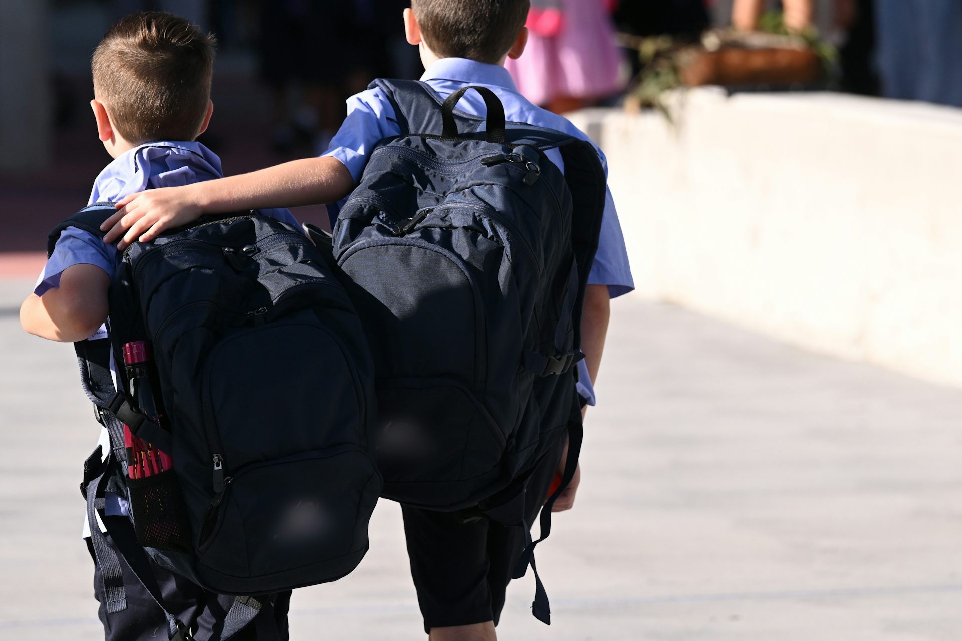 Two primary students walk with backpacks. One puts their arm around the other.