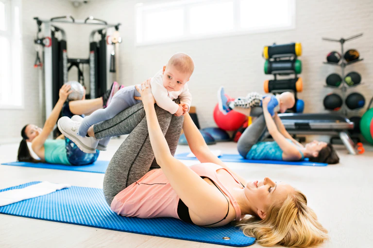 Women in a gym lie on mats doing stretches using their babies as weights.