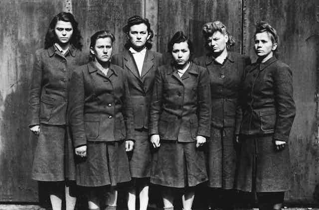 Six female SS guards from Belsen standing in a row scowling at the camera.