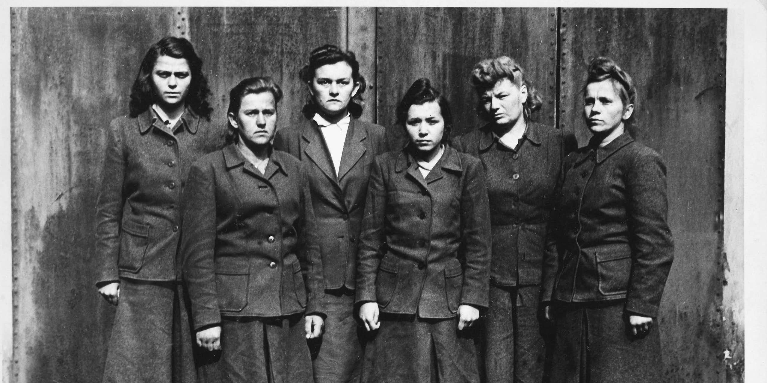 Six female SS guards from Belsen standing in a row scowling at the camera.