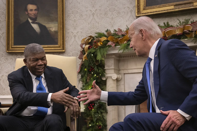 Two smiling men wearing suits reach out to shake hands while seated.