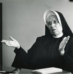 A woman in a black and white nun's habit gestures while speaking in a room with white walls.