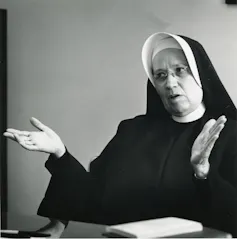 A woman in a black and white nun's habit gestures while speaking in a room with white walls.