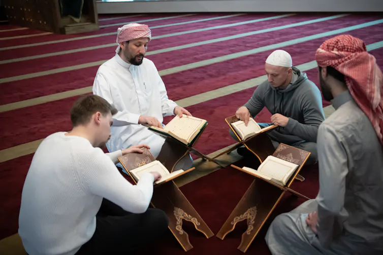 Four men, two of whom wear a robe-like garment, sit on a red carpet studying books in small stands.