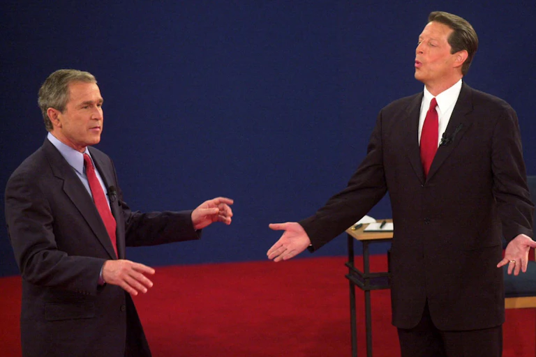 Two men in suits make gestures while standing on a stage with a red carpet and blue background.