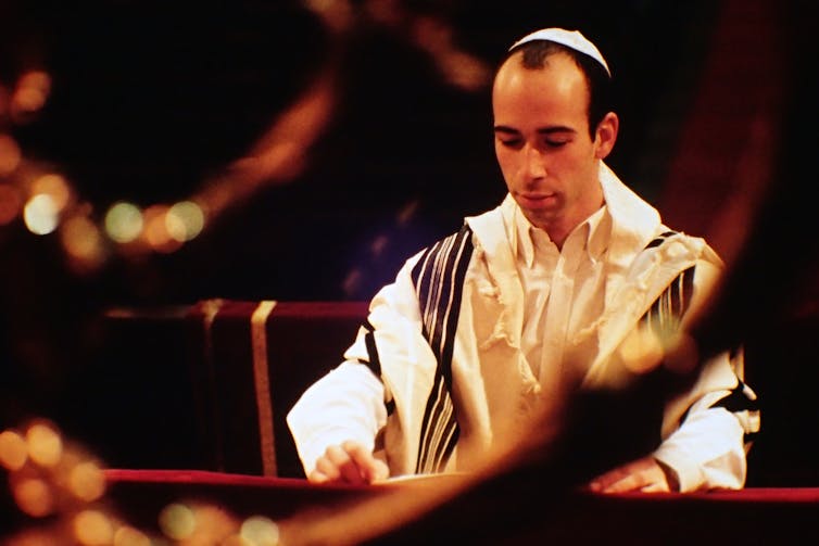 A man in a blue and white prayer robe and white cap stands as he looks down at a text.