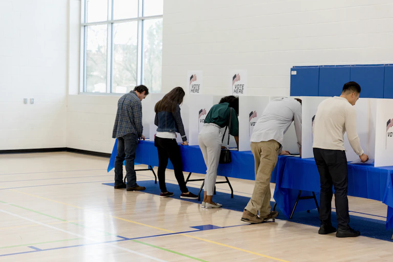 Five people standing at voting booths in a large room.