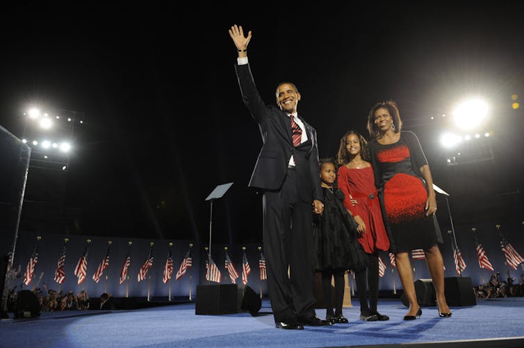 A man in a dark suit waving from a stage in the night with a woman and two girls next to him.