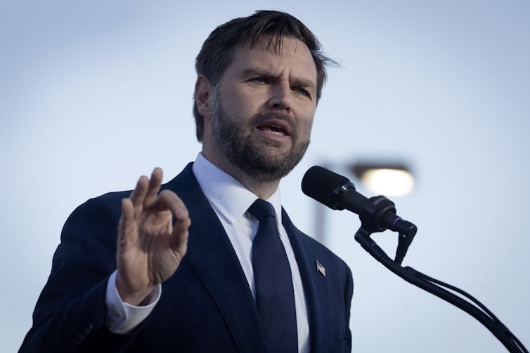 A bearded man in a blue suit and tie talking into a microphone.