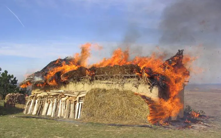 primitive building on fire with wood and straw piled up next to the wall
