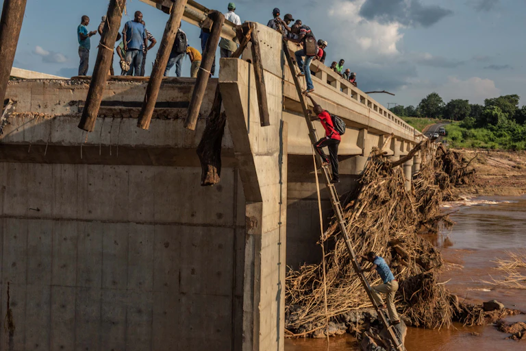 People climb a ladder to cross a large bridge that has been partially destroyed. Other people stand a top the bridge.