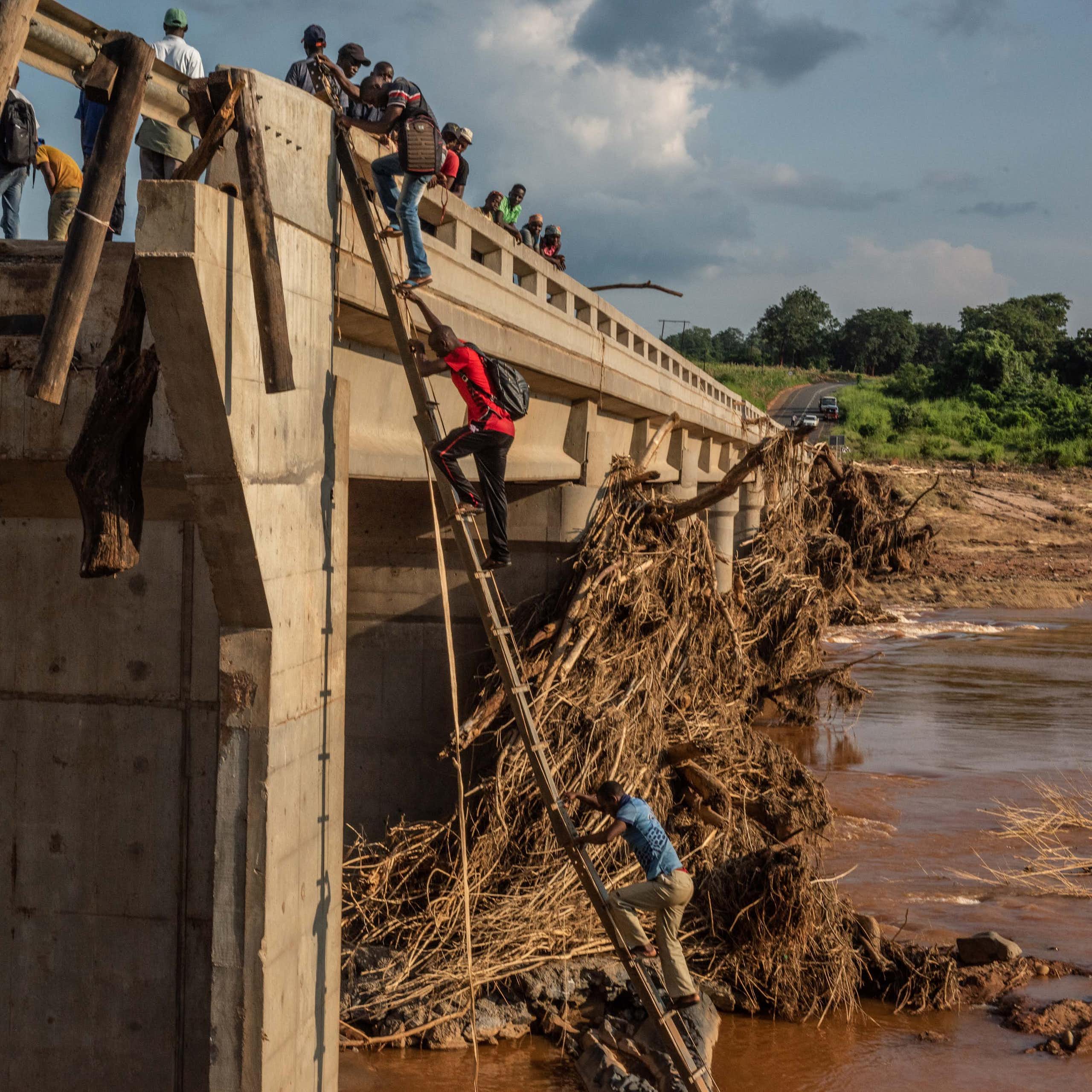 People climb a ladder to cross a large bridge that has been partially destroyed. Other people stand a top the bridge.
